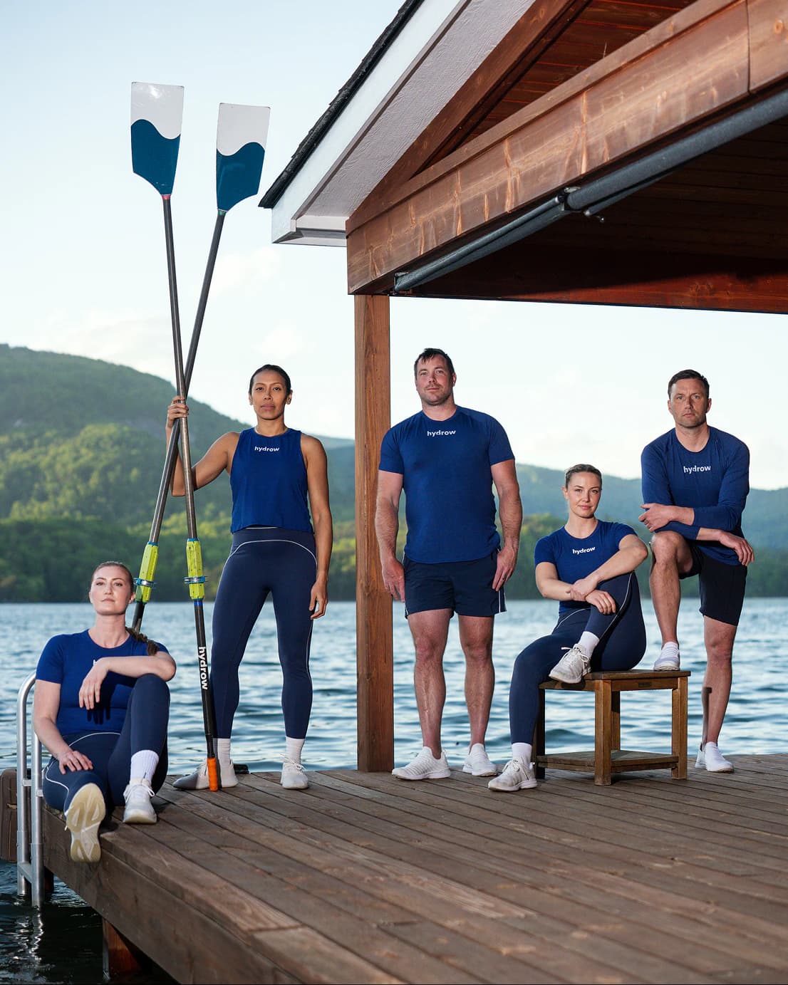 Hydrow athletes standing together on a lakeside dock holding oars