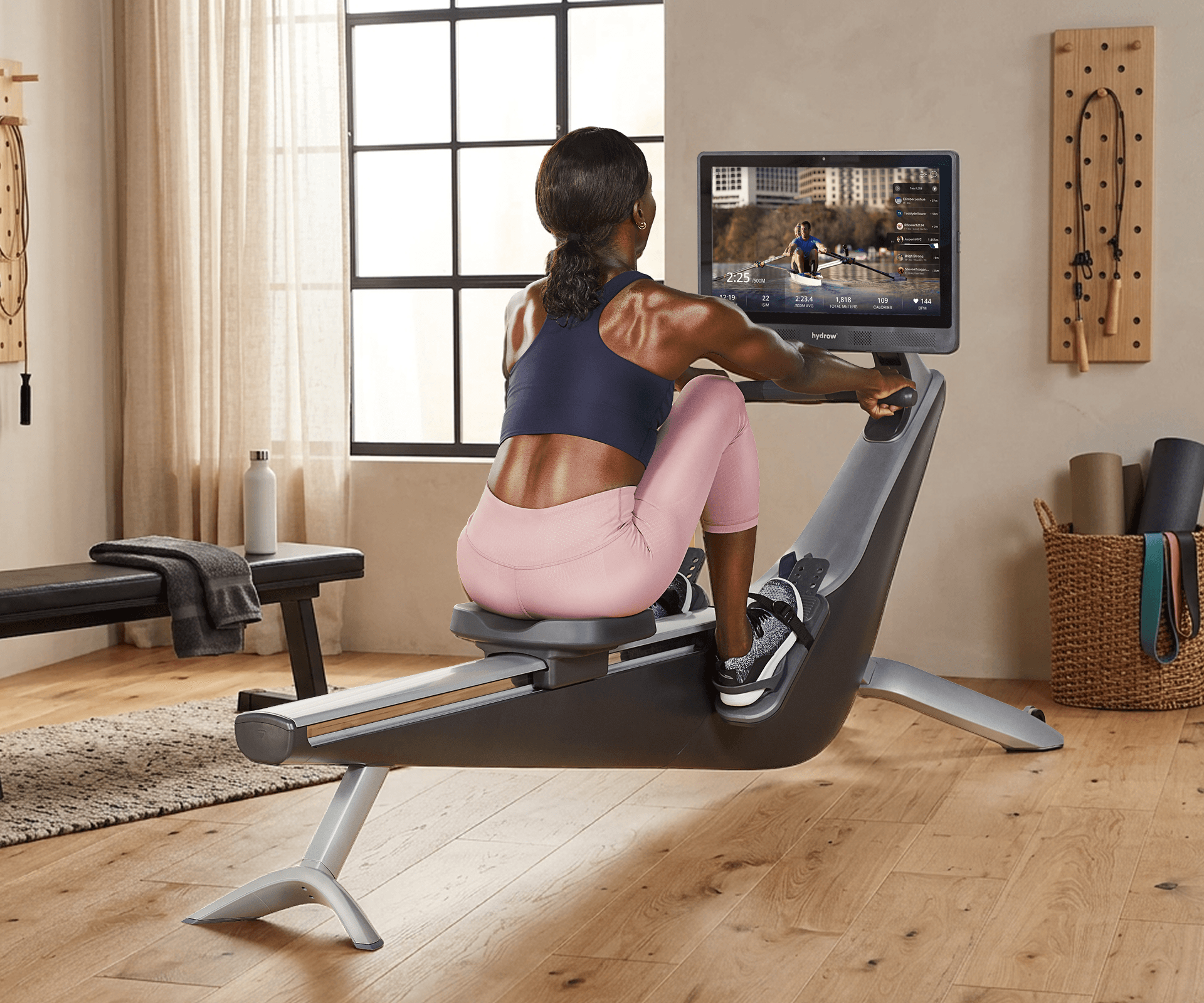 Woman exercising on a Hydrow Origin rower, watching a virtual rowing workout on screen.