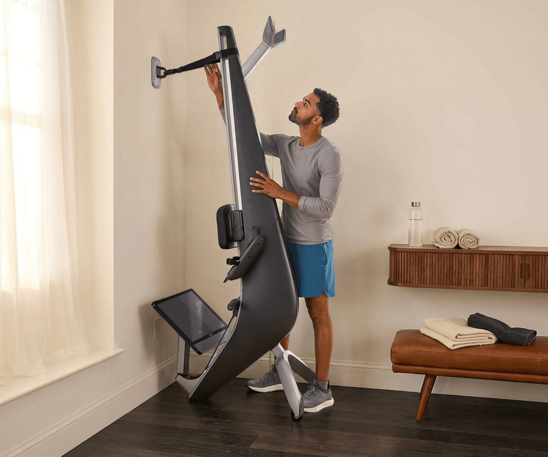 Man securing a Hydrow Arc rower into vertical storage against a wall.