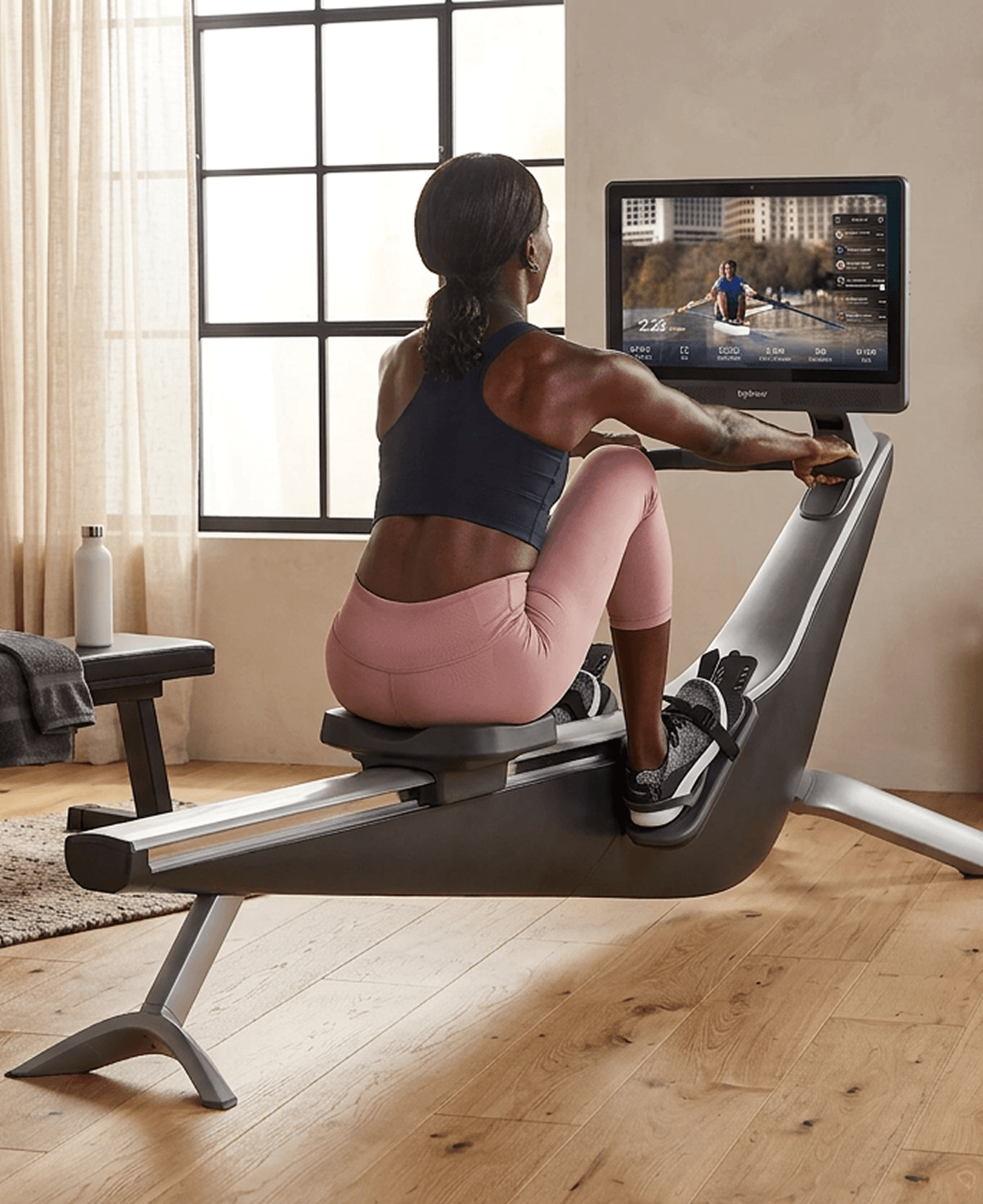 A person rowing on a Hydrow Origin rower in a sunlit room while following an on-water class on the monitor