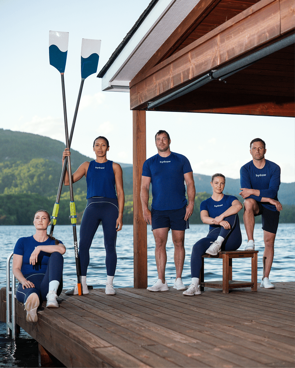 Hydrow athletes standing together on a lakeside dock holding oars