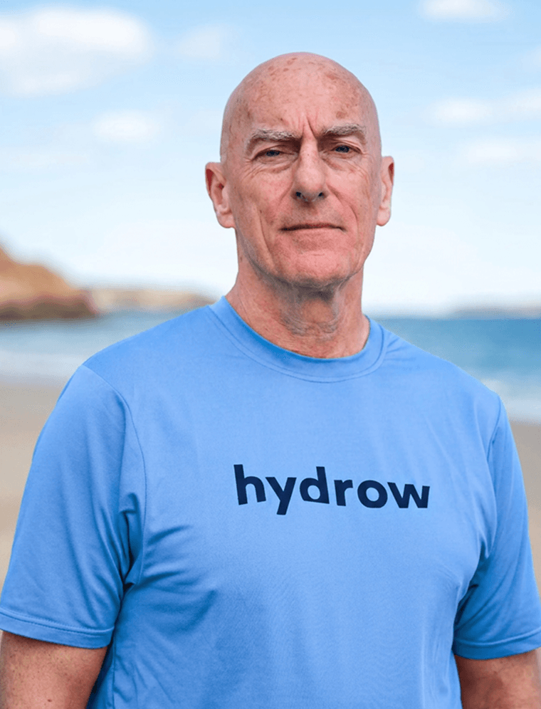 Hydrow athlete Peter Donohoe standing outdoors in a blue Hydrow shirt by the beach