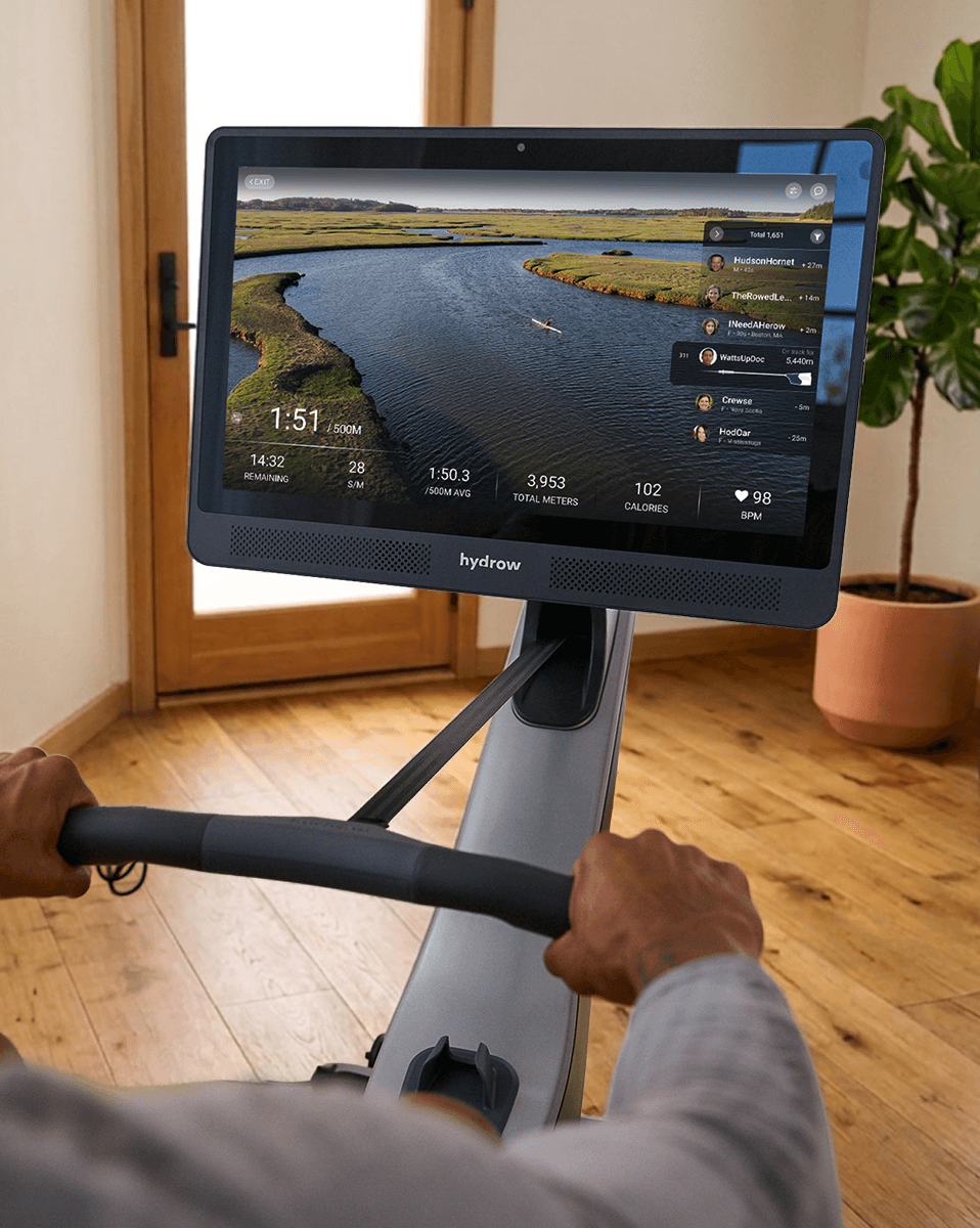 A Hydrow rower mid-stroke with a scenic waterway on the screen