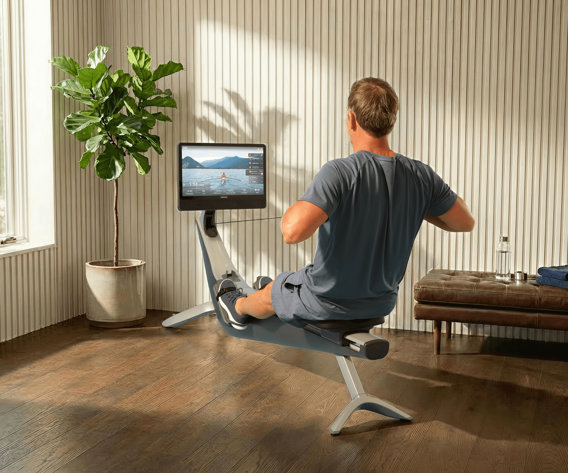 Man rowing on a Hydrow Arc, watching a live outdoor rowing workout on screen.