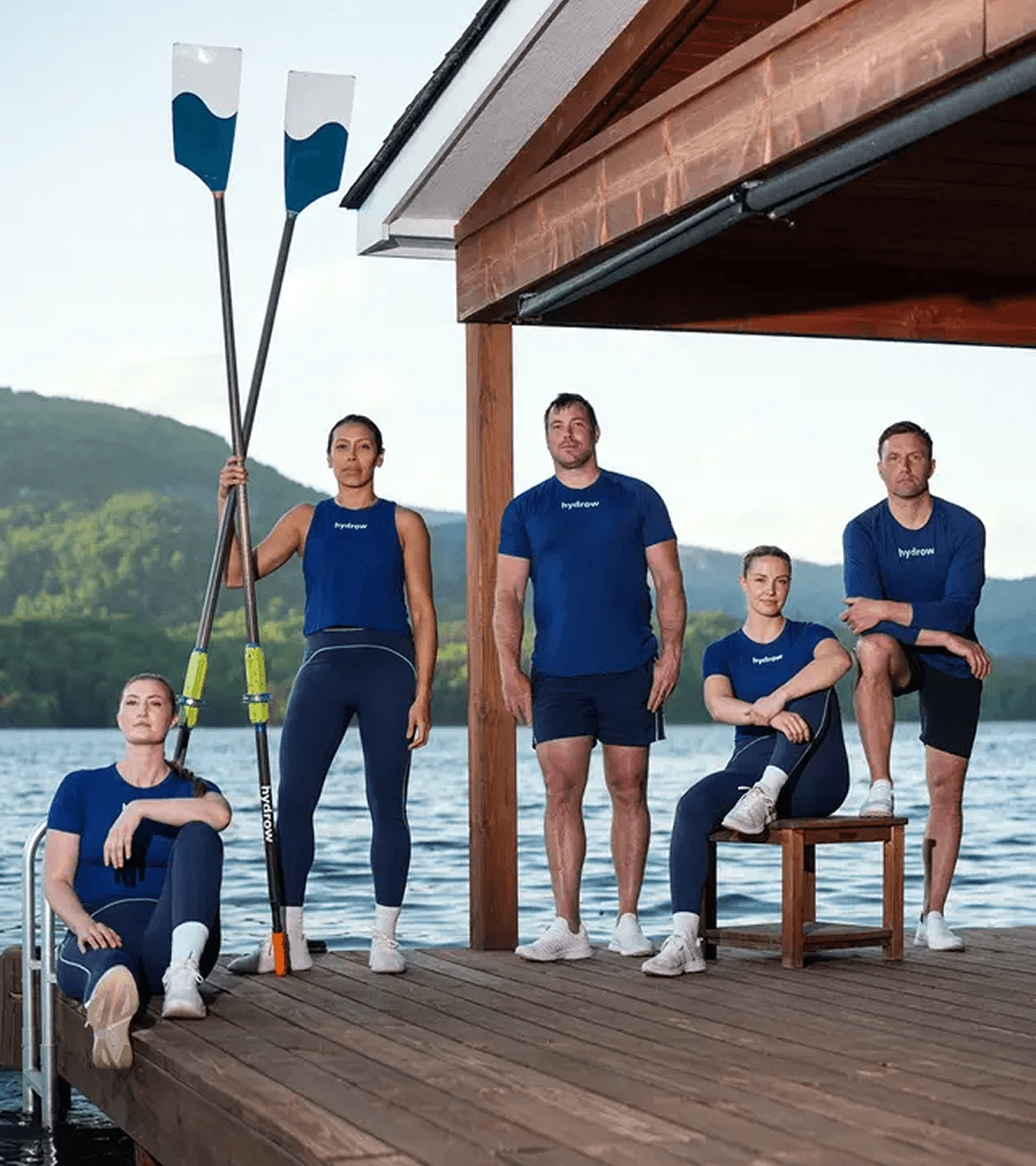 Hydrow athletes standing together on a lakeside dock holding oars